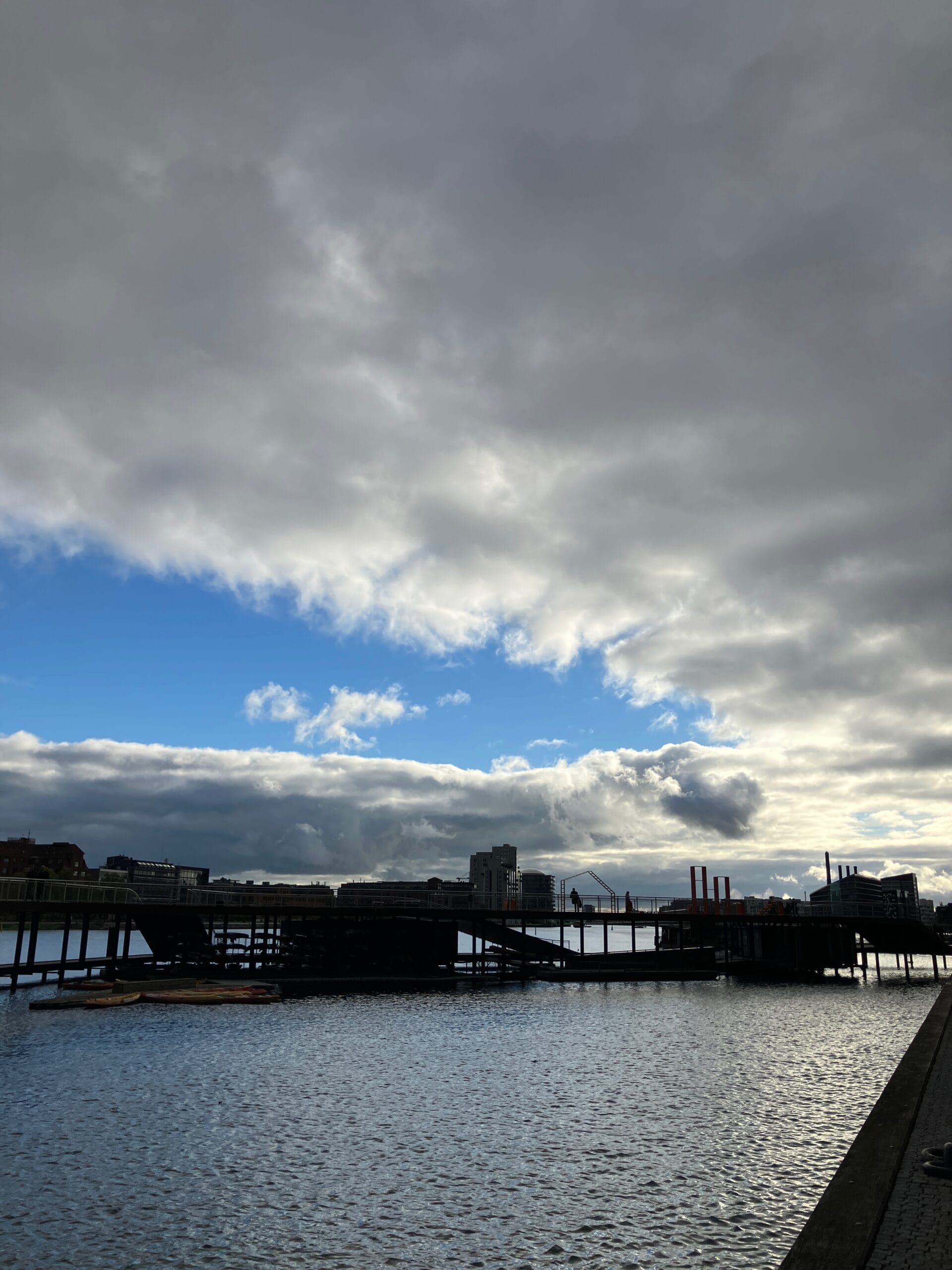 Een bewolkte donkergrijze hemel met een blauwe streep. In het water is een houten constructie waar mensen op lopen