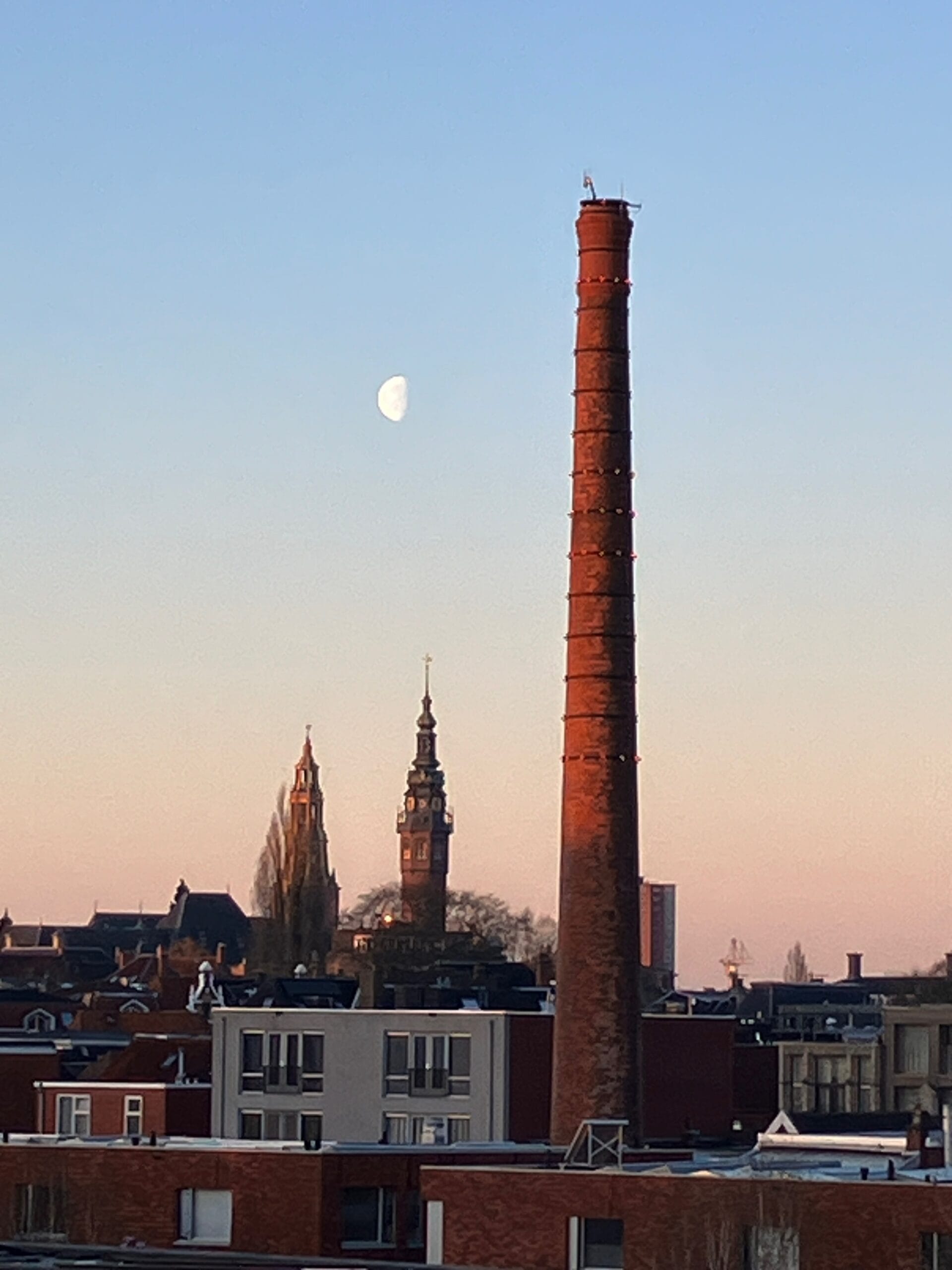 Schoorsteen en twee kerktoren voor een avondhemel met een halve maan aan de hemel